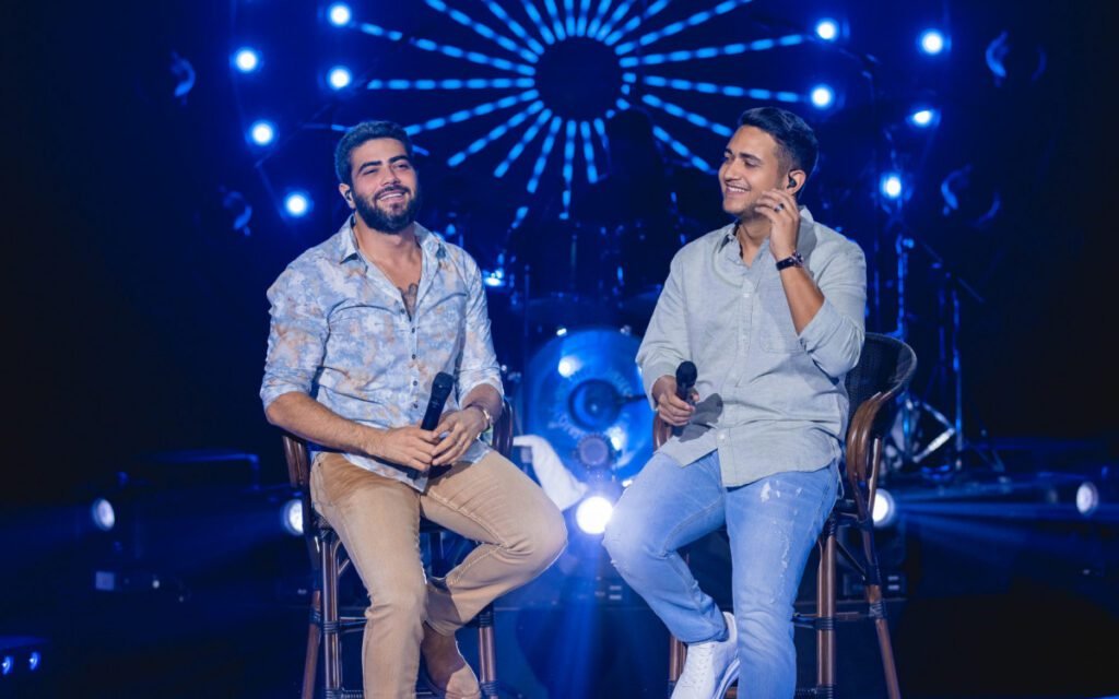 Henrique e Juliano cantando abraçados no palco durante a turnê Manifesto Musical em 2026, com luzes de estádio ao fundo.