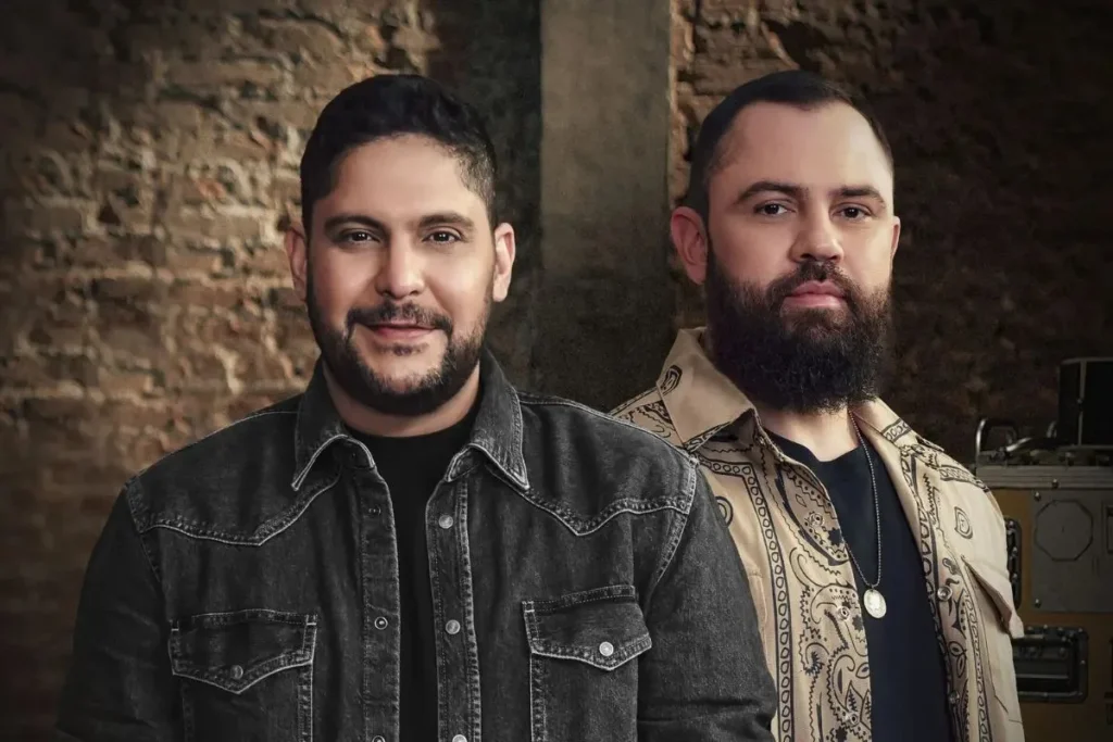 orge e Mateus cantando juntos no palco durante a turnê de 20 anos em 2026, com luzes brilhantes e multidão ao fundo.